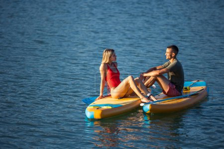 Téléchargez les photos : Jeune et beau couple d'amoureux assis sur une planche de surf en pleine mer. - en image libre de droit