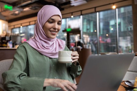 Foto de Nómada digital. Mujer joven en hijab trabajando en una cafetería y buscando contenta - Imagen libre de derechos