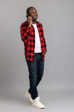 Photo for Happy black bearded man wearing red checkered shirt talking on smartphone posing isolated over gray studio background. - Royalty Free Image