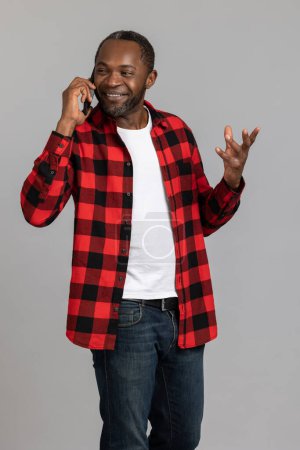 Foto de Hombre barbudo negro feliz con camisa a cuadros roja hablando en el teléfono inteligente posando aislado sobre fondo gris estudio. - Imagen libre de derechos