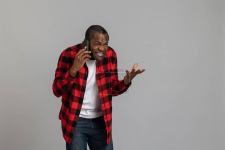 Photo for Aggressive angry black bearded man wearing red checkered shirt screaming while talking phone posing isolated over gray studio background. - Royalty Free Image