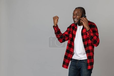 Photo for Overjoyed black bearded man wearing red checkered shirt talking phone clenched fist hearing good news posing isolated over gray studio background. - Royalty Free Image