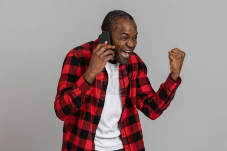 Foto de Extremadamente feliz negro barbudo hombre usando rojo a cuadros camisa hablando teléfono apretado puño posando aislado sobre gris estudio fondo. - Imagen libre de derechos