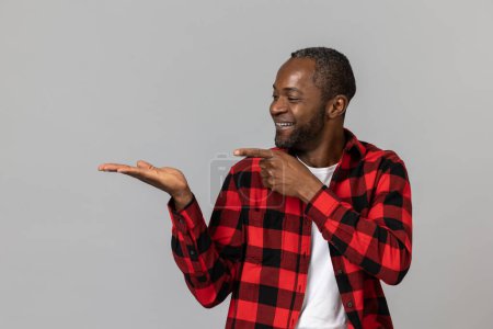 Téléchargez les photos : Joyeux homme barbu noir portant chemise à carreaux rouge présentant l'espace de copie sur la paume posant isolé sur fond de studio gris. - en image libre de droit