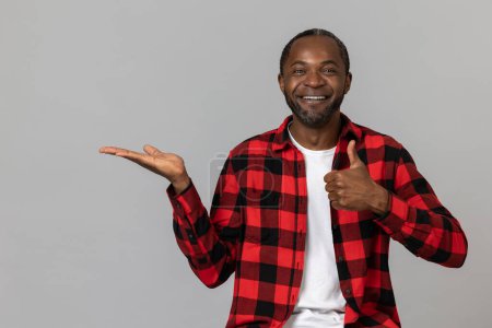 Photo for Joyful black bearded man wearing red checkered shirt presenting copy space on palm showing thumb up posing isolated over gray studio background. - Royalty Free Image