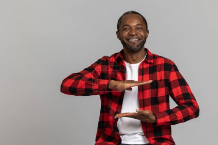 Photo for Cheerful black bearded man wearing red checkered shirt showing empty space between hands posing isolated over gray studio background. - Royalty Free Image