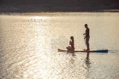 Téléchargez les photos : Femme méditant sur supboard tôt le matin avec l'homme pagayant au coucher du soleil. - en image libre de droit