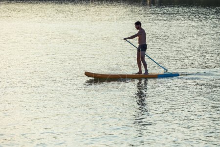 Téléchargez les photos : Homme debout sur le supboard au milieu du lac au coucher du soleil. - en image libre de droit