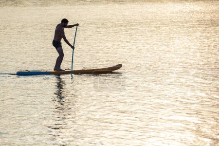 Téléchargez les photos : Homme debout sur le supboard au milieu du lac au coucher du soleil. - en image libre de droit