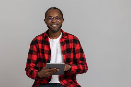 Photo for Happy black bearded man wearing red checkered shirt and glasses using digital tablet posing isolated over gray studio background. - Royalty Free Image