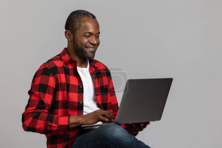 Foto de Agradable hombre barbudo negro con camisa a cuadros roja usando el ordenador portátil para el trabajo remoto posando aislado sobre fondo de estudio gris. - Imagen libre de derechos