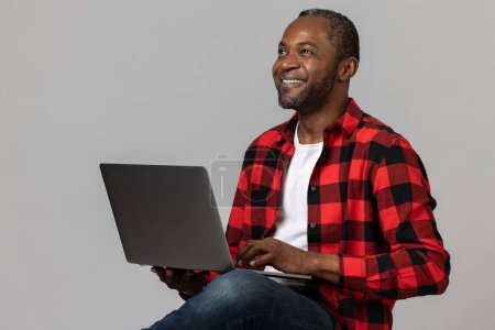 Photo for Pleased black bearded man wearing red checkered shirt using laptop for remote work posing isolated over gray studio background. - Royalty Free Image