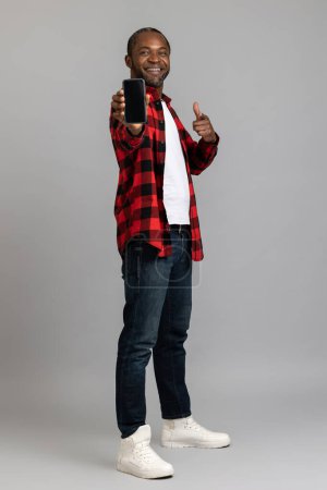 Photo for Joyful black bearded man wearing red checkered shirt showing smartphone with empty screen posing isolated over gray studio background. - Royalty Free Image