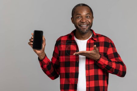 Foto de Hombre barbudo negro satisfecho con camisa a cuadros roja que muestra el teléfono móvil con pantalla en blanco posando aislado sobre fondo de estudio gris. - Imagen libre de derechos
