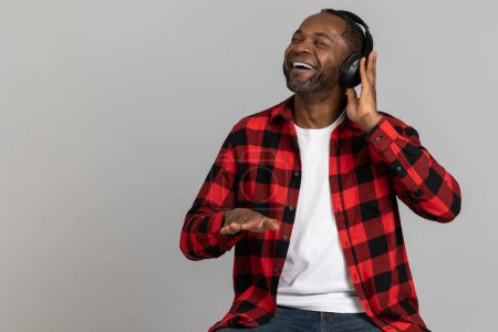 Photo for Satisfied black bearded man dj in headphones wearing red checkered shirt enjoying music posing isolated over gray studio background. - Royalty Free Image