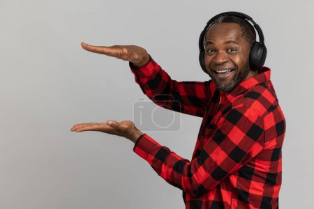 Photo for Cheerful black bearded man in headphones wearing red checkered shirt showing advertisement area posing isolated over gray studio background. - Royalty Free Image