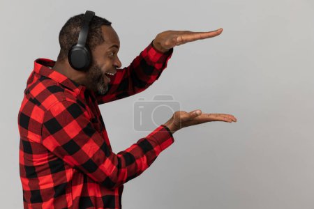 Foto de Encantado hombre barbudo negro en auriculares con camisa a cuadros roja que muestra el área de publicidad posando aislado sobre fondo gris estudio. - Imagen libre de derechos