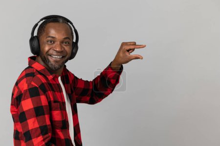 Foto de Hombre barbudo negro feliz en auriculares con camisa a cuadros roja que muestra un tamaño pequeño posando aislado sobre fondo gris del estudio. - Imagen libre de derechos