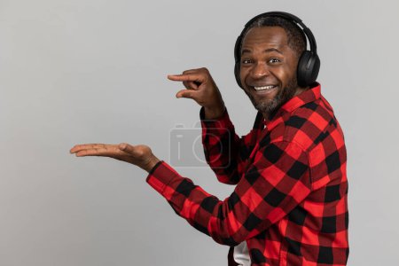 Foto de Alegre hombre barbudo negro en auriculares con camisa a cuadros roja que muestra tamaño pequeño y área de publicidad en la palma posando aislado sobre fondo gris estudio. - Imagen libre de derechos