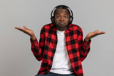 Photo for Uncertain puzzled black bearded man in headphones wearing red checkered shirt posing isolated over gray studio background. - Royalty Free Image