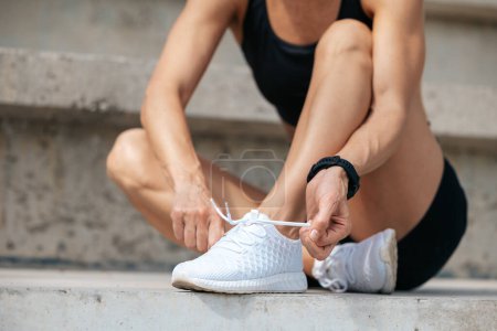 Photo for Female runner kneeling and tying shoelace on street on sunny day. - Royalty Free Image