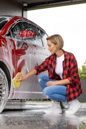 Foto de Lavado de coches. Mujer rubia limpiando la puerta del coche con una espuma - Imagen libre de derechos