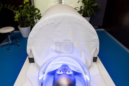 Foto de Mujer en gafas protectoras acostada bajo lámpara led para la terapia de luz de color en el salón de belleza. - Imagen libre de derechos