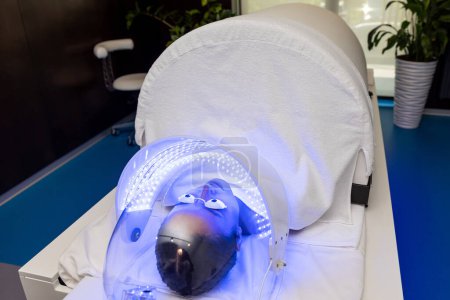 Foto de Mujer en gafas protectoras acostada bajo lámpara led para la terapia de luz de color en el salón de belleza. - Imagen libre de derechos