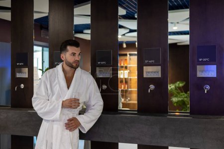 Téléchargez les photos : Bel homme caucasien en peignoir blanc eau potable, profiter de sa journée de congé au luxueux centre de spa de bien-être. - en image libre de droit