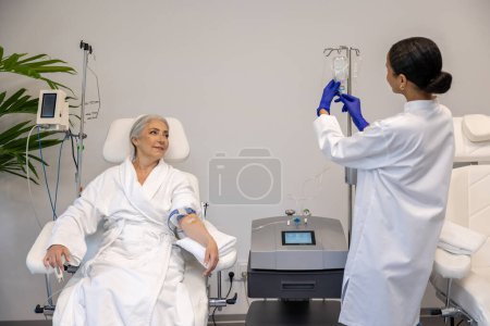 Foto de Mujer mayor que recibe terapia con vitaminas intravenosas en la habitación del hospital sentada en un sillón unido al goteo de infusión de vitamina IV en el salón de belleza. - Imagen libre de derechos