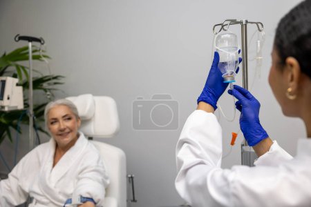 Photo for Mature woman receiving intravenous vitamin therapy in hospital room sitting in armchair attached to vitamin IV infusion drip in wellness center or beauty salon. - Royalty Free Image