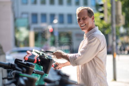 Téléchargez les photos : Sourire mec d'âge moyen en utilisant un smartphone pour débloquer scooter électrique partagé dans la rue de la ville. - en image libre de droit