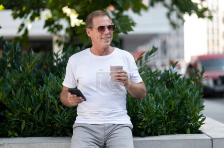 Foto de Guapo hombre caucásico de mediana edad usando teléfono inteligente beber café al aire libre en el parque de la ciudad. - Imagen libre de derechos