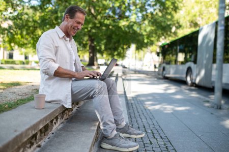 Foto de Vista lateral del programador freelancer de mediana edad sentado en el banco trabajando en línea en el portátil en el parque de la ciudad. - Imagen libre de derechos
