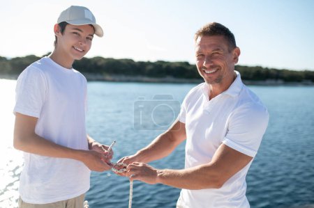 Téléchargez les photos : Bonne famille. Homme mûr et son fils passer du temps sur le yacht et regarder heureux - en image libre de droit