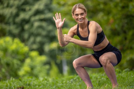 Photo for Working out. Squatting practice. Beautiful woman working out doing squats in summer park, waving hand greeting somebody. - Royalty Free Image