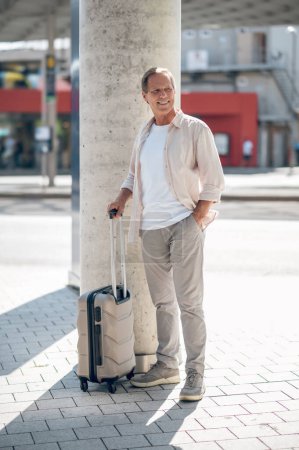 Foto de Sonriente hombre de pie con la maleta en la calle de la ciudad. - Imagen libre de derechos