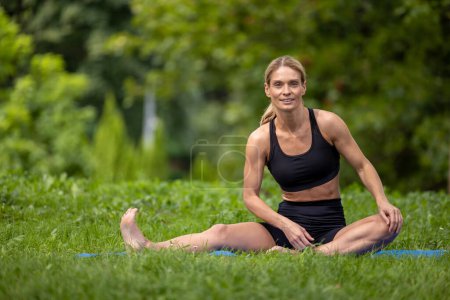 Foto de Balance de bienestar. Práctica de relajación. Aptitud activa. Mujer estirando el cuerpo sentado en la alfombra de ejercicio en el parque verde. - Imagen libre de derechos