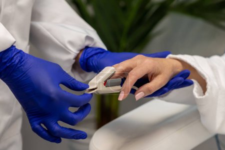 Téléchargez les photos : Infirmière en gants bleus vérifiant le niveau d'oxygène au patient féminin dans le salon de beauté avant les procédures de cosmétologie. - en image libre de droit