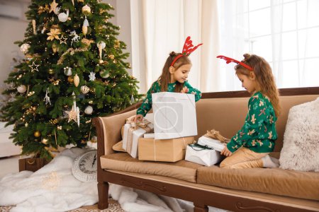 Foto de Dos hermanas en jerséis verdes y cuernos de ciervo abren regalos de Navidad - Imagen libre de derechos