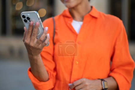Téléchargez les photos : Femme méconnaissable portant une chemise orange debout en plein air avec smartphone dans les mains. - en image libre de droit