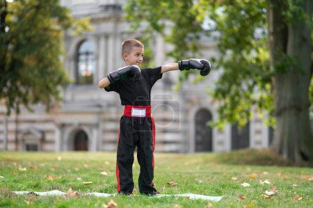 Téléchargez les photos : La boxe. Enfant blond en gants de boxe faisant de l'exercice dans le parc - en image libre de droit