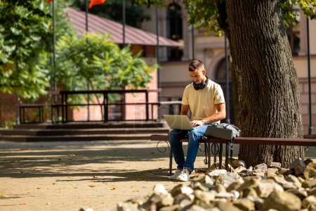 Photo for Studying. Short-haired young guy sitting under the tree and working on a laptop - Royalty Free Image