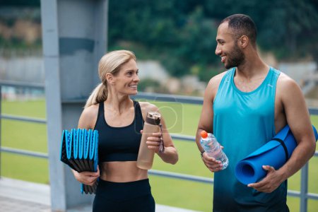Foto de Hermosa pareja de fitness con ropa deportiva elegante posando con esterilla de yoga y botellas de agua antes de entrenar en el estadio. - Imagen libre de derechos