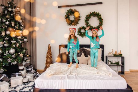 Photo for Cute twins in pajamas and santa hats jumping on bed - Royalty Free Image