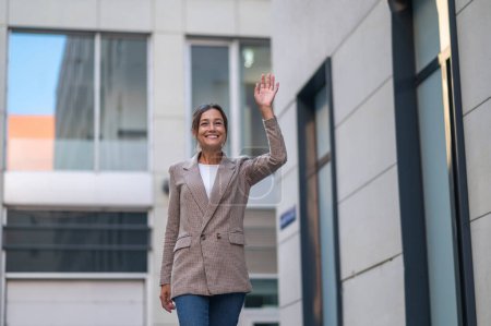 Photo for Brunette woman wearing beige suit posing near urban building waving hand greeting somebody. - Royalty Free Image