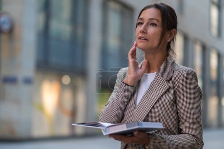 Téléchargez les photos : Femme d'affaires confiante pensive portant une veste beige tenant le livre tout en posant sur la rue de la ville. - en image libre de droit
