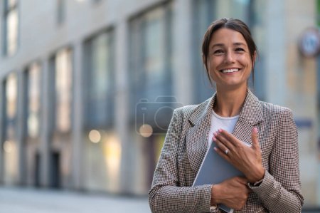 Photo for Smiling business woman wearing beige jacket holding book while posing on city street. - Royalty Free Image