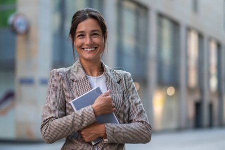 Téléchargez les photos : Plan extérieur d'une femme heureuse tenant son planificateur ou son livre, vêtue d'une veste beige. - en image libre de droit