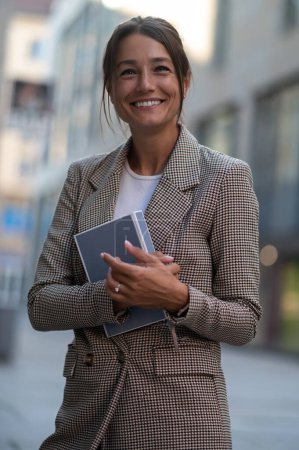 Foto de Foto al aire libre de una mujer sonriente sosteniendo su planificador o libro, usando chaqueta beige. - Imagen libre de derechos